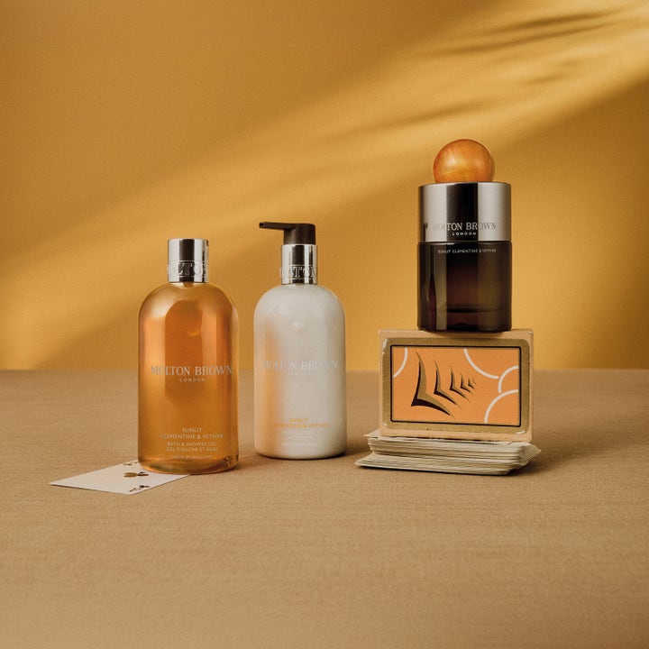 Molton Brown Sunlit Clementine & Vetiver 300 ml dusjsåpe i en klar flaske i et varmt, gult miljø med spillkort.