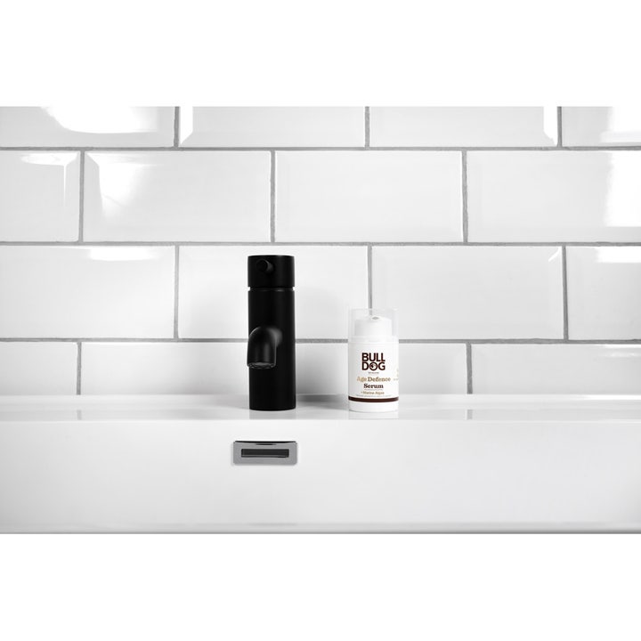 Bulldog Age Defence Serum 50 ml i hvit pumpeflaske står ved siden av en sort kran i et flislagt baderomsmiljø.
