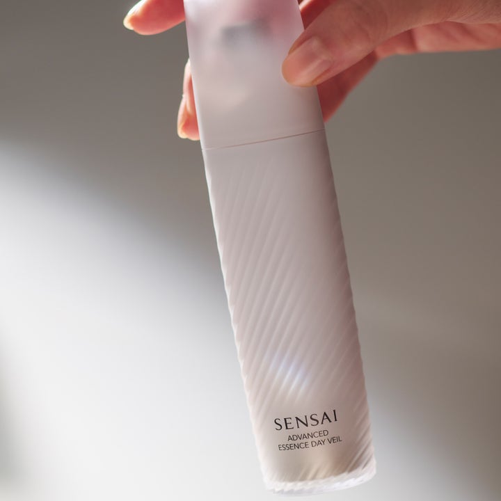 Sensai Advanced Essence Day Veil 40 ml fuktighetsgivende serum i frostet hvit flaske med riller holdt av en hånd.