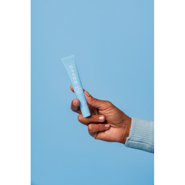 MASHH Bright Eye Cream 15 ml fuktighetsgivende øyekrem i lyseblå tube holdt av en hånd mot en blå bakgrunn.