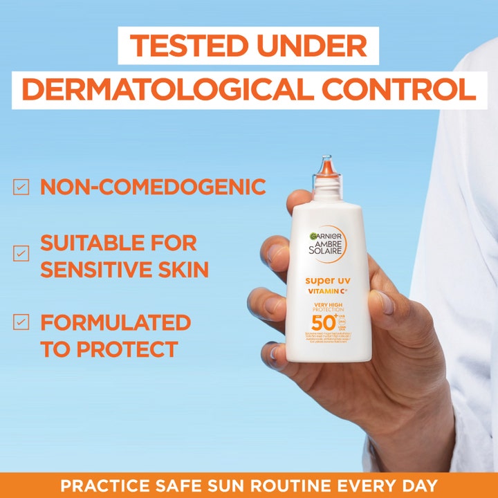 Garnier Ambre Solaire Super UV Vitamin C 40 ml solkrem mot mørke flekker i hvit flaske holdt av en hånd mot blå bakgrunn.