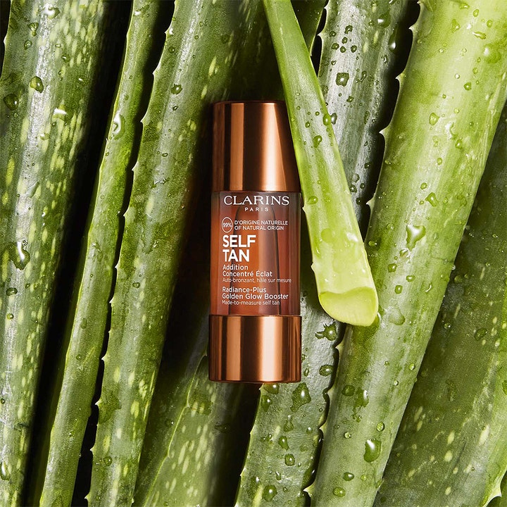 Clarins Radiance-Plus Golden Glow Booster Face 15 ml selvbruning i bronseflaske omgitt av fuktige aloe vera-blader.
