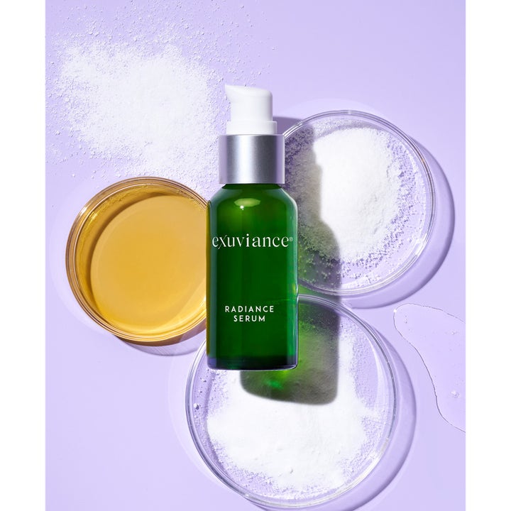 Exuviance Radiance Serum 30 ml glødgivende serum i grønn glassflaske med ingredienser i petriskåler på lilla bakgrunn