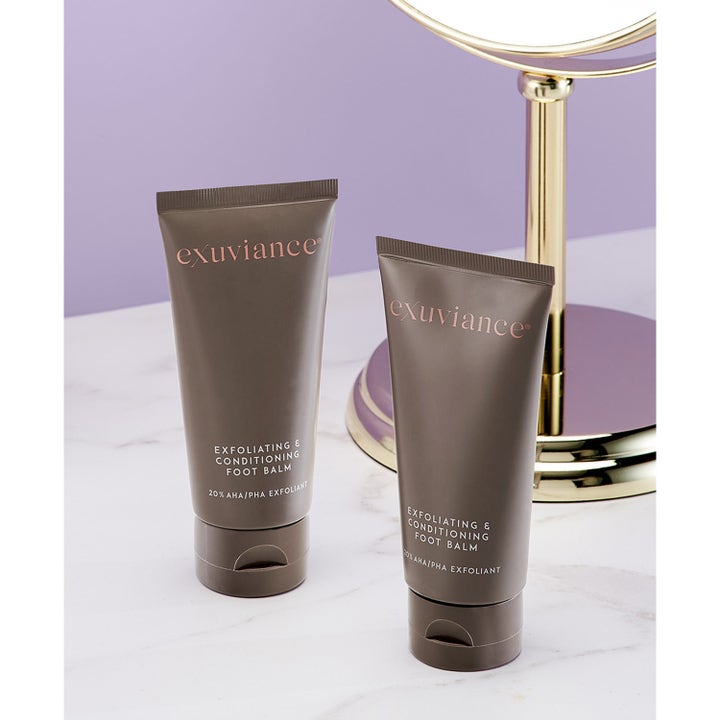 Exuviance Exfoliating & Conditioning Foot Balm 50 g eksfolierende fotkrem i brune tuber på marmor ved et gullspeil.