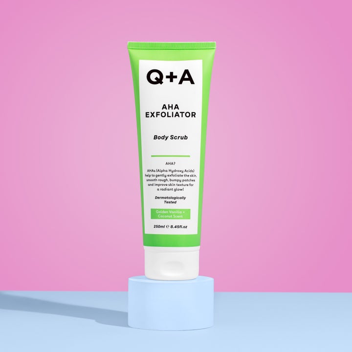 Q+A AHA Exfoliator Body Scrub 250 ml eksfolierende kroppsskrubb i grønn tube på lyseblå sokkel med rosa bakgrunn.