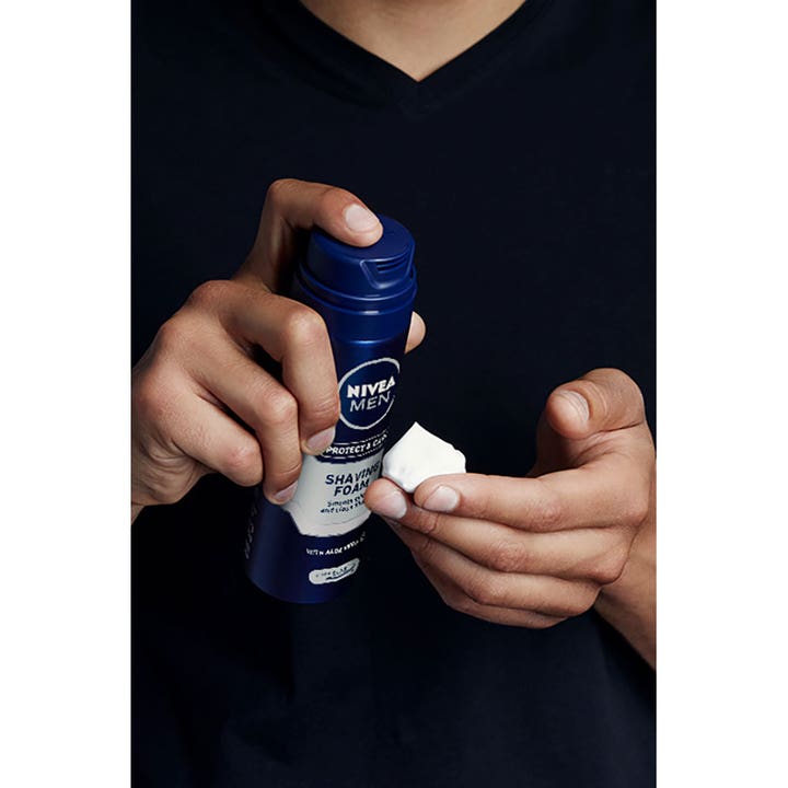 NIVEA Protect & Care Shaving Foam 200 ml pleiende barberskum i blå boks som sprayes som hvitt skum i en hånd.