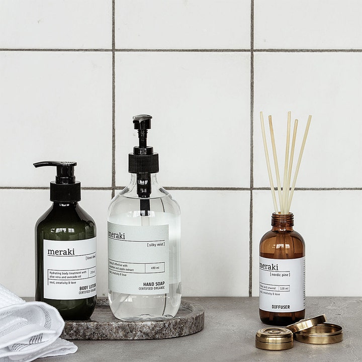 Meraki Linen Dew Body Lotion 275 ml fuktighetsgivende hudpleie i mørk pumpeflaske ved siden av duftpinner