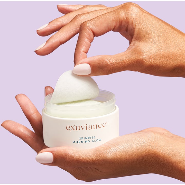 Exuviance SkinRise Morning Glow 36 stk serum pads som gir glød i hvit krukke holdt av hender mot lilla bakgrunn