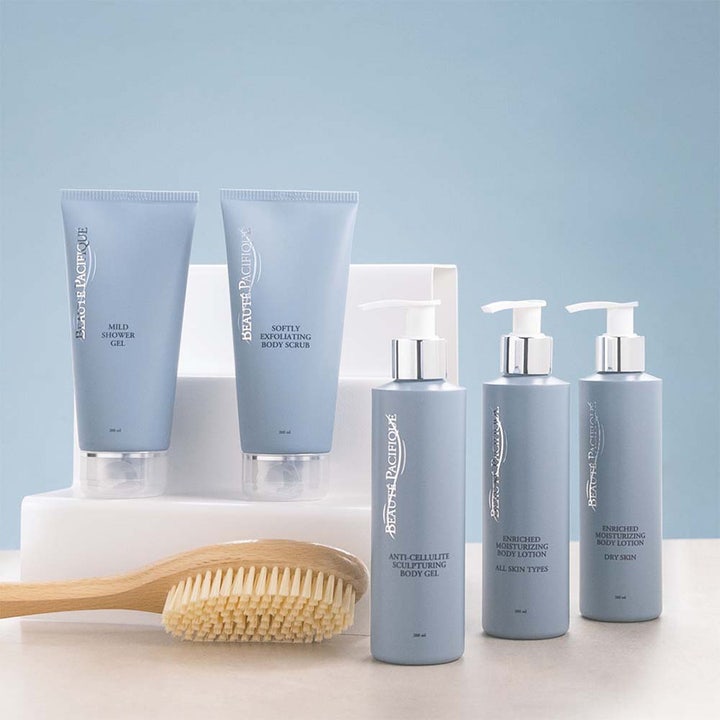 Beauté Pacifique kroppspleie sett med dusjgelé, skrubb og body lotion i blå tuber og flasker ved en badebørste.