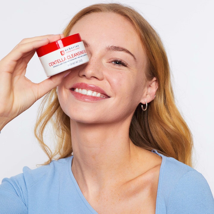 Erborian Centella Cleansing Balm 80 g ansiktsrengjøring i hvit krukke holdt av en smilende kvinne