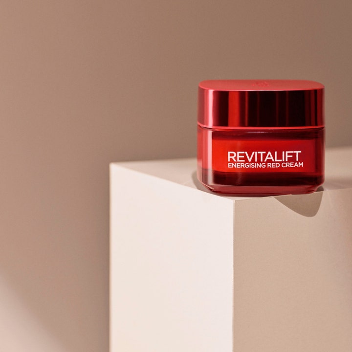 L'Oréal Paris Revitalift Ginseng Glow Day Cream 50 ml glødgivende dagkrem i rød glasskrukke på en beige blokk