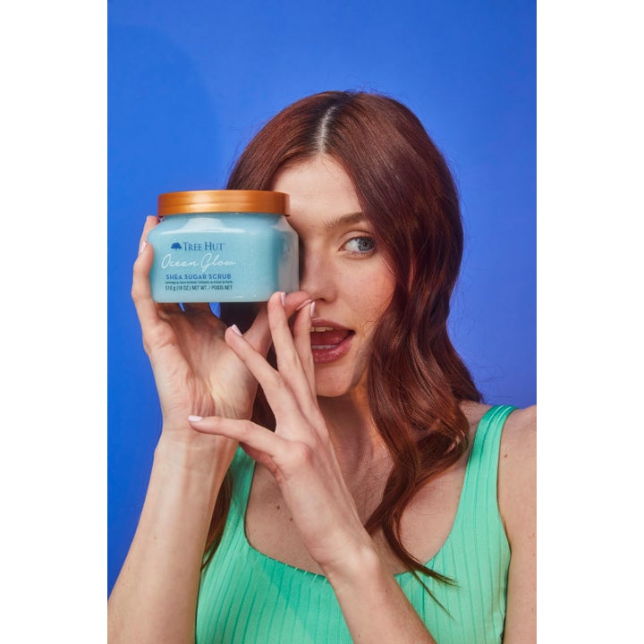 Tree Hut Shea Sugar Scrub Ocean Glow 510 g kroppsskrubb kvinne holder lyseblå krukke med gullokk foran ansiktet.