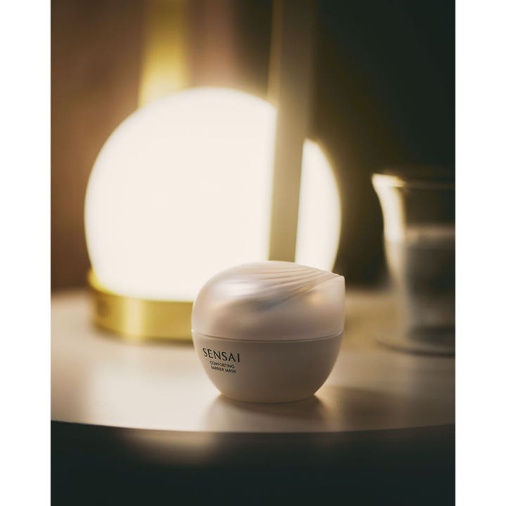 Sensai Comforting Barrier Mask 60 ml styrkende ansiktsmaske i hvit krukke ved en stemningsfull glødende lampe.