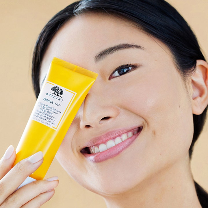 Origins Drink Up 10 Minute Mask 75 ml fuktighetsgivende ansiktsmaske i gul tube holdt av en smilende kvinne
