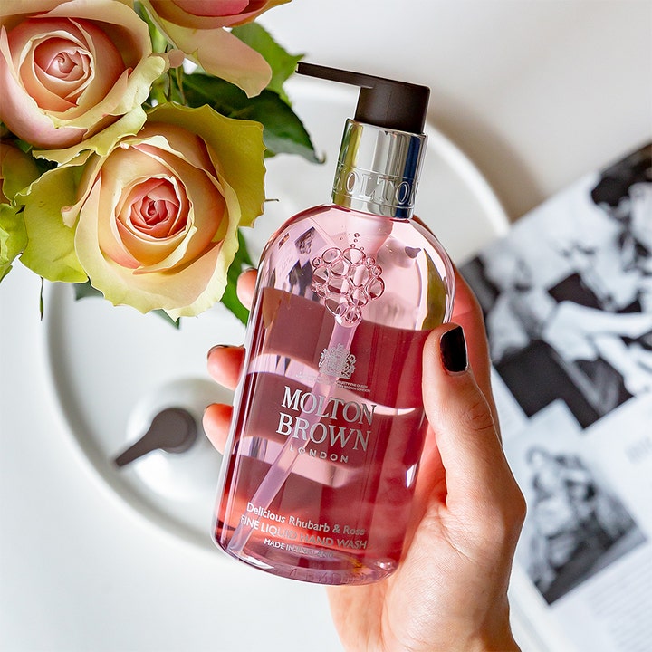 Molton Brown Delicious Rhubarb & Rose 300 ml flytende håndsåpe i klar flaske holdt i en hånd ved siden av friske roser.