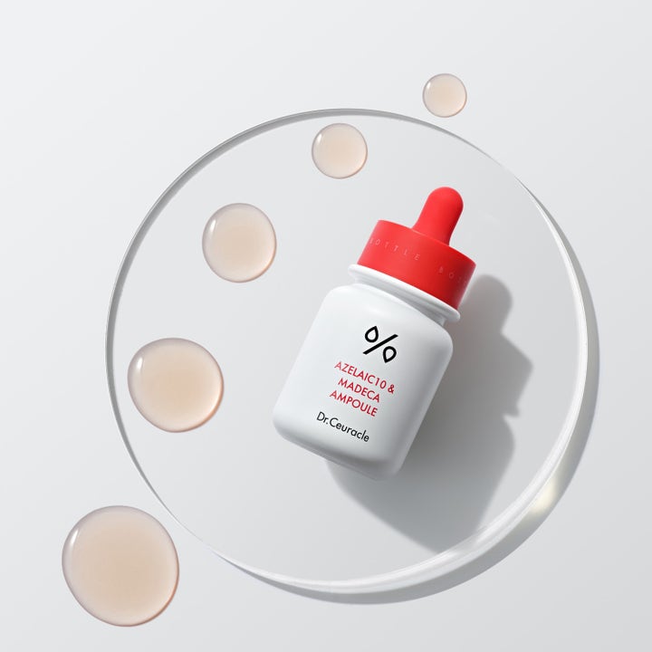 Dr. Ceuracle Azelaic 10 & Madeca Ampoule 30 ml serum i hvit flaske med rød kork liggende ved siden av klare dråper.