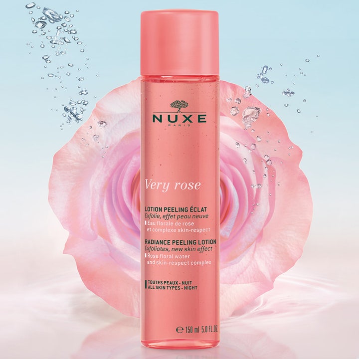 Nuxe Very Rose Peeling Lotion 150 ml eksfolierende peeling i rosa flaske foran en stor rose som gir glød.