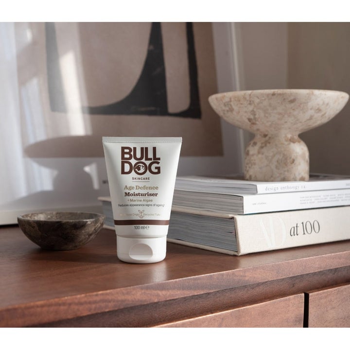 Bulldog Age Defence Moisturiser 100 ml fuktighetskrem mot rynker i hvit tube på en treflate ved siden av bøker.