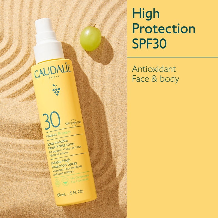 Caudalie Vinosun High Protection Spray 150 ml solkrem med SPF30 i gul flaske liggende i sanden ved en grønn drue.