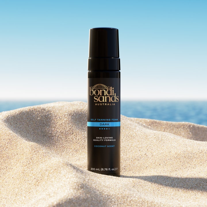 Bondi Sands Self Tanning Foam Dark 200 ml gir dyp glød i svart pumpeflaske plassert i sand ved stranden.