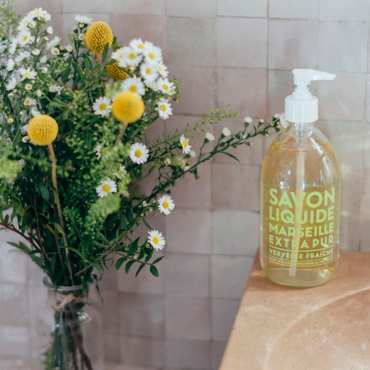 Compagnie de Provence Liquid Marseille Soap Fresh Verbena 495 ml i glassflaske ved siden av en blomsterbukett.