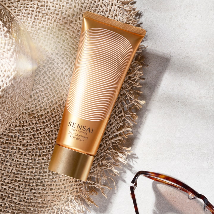 Sensai Silky Bronze Self Tanning For Body 150 ml gir glød i gullfarget tube på en stråhatt med solbriller.