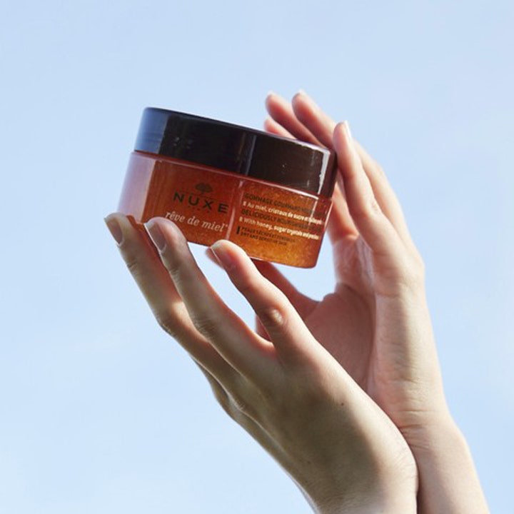 Nuxe Rêve de Miel Body Scrub 175 ml eksfolierende kroppsskrubb i ravfarget krukke holdt mot en blå himmel