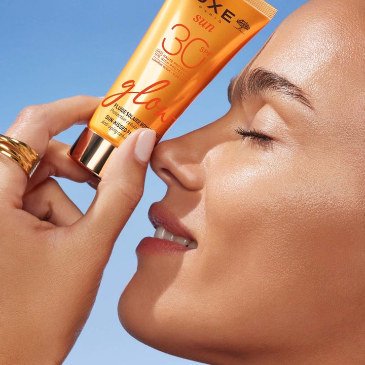 Nuxe Sun-Kissed Fluid 40 ml solkrem for ansikt gir huden glød vist med oransje tube og kvinne mot blå himmel