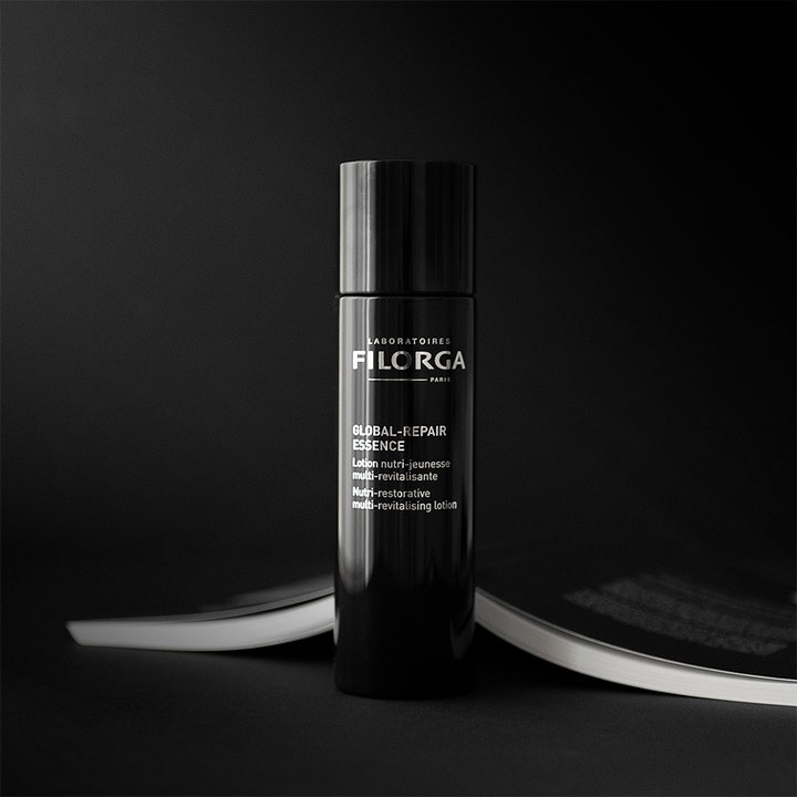 Filorga Global-Repair Essence 150 ml revitaliserende ansiktsvann i elegant svart flaske stående ved en åpen bok