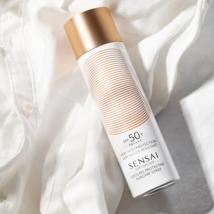 Sensai Silky Bronze Cooling Protective Suncare Spray 150 ml solkrem gir SPF 50 i hvit sprayflaske med gulllokk på hvit skjorte.