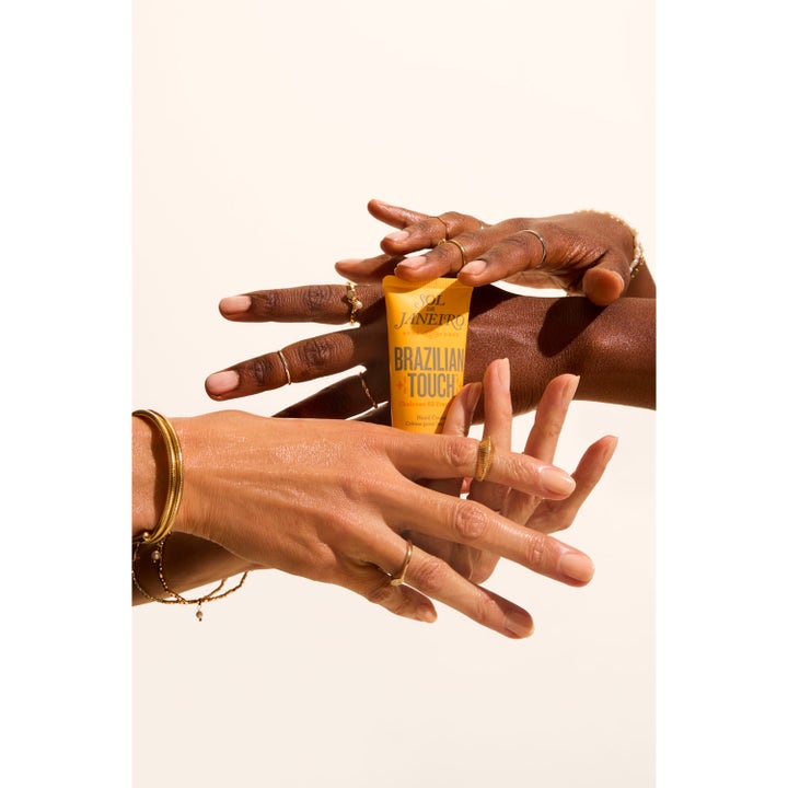 Sol de Janeiro Brazilian Touch Hand Cream 50 ml fuktighetsgivende handkrem i gul tube holdt av hender med gullsmykker.