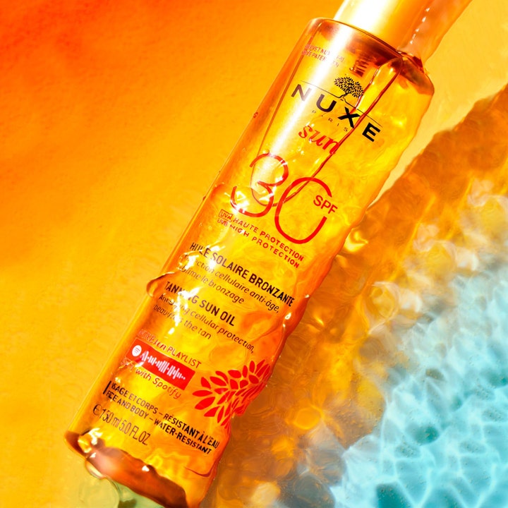 Nuxe Sun Tanning Oil 150 ml sololje SPF30 gir glød i en oransje sprayflaske nedsenket i glitrende vann