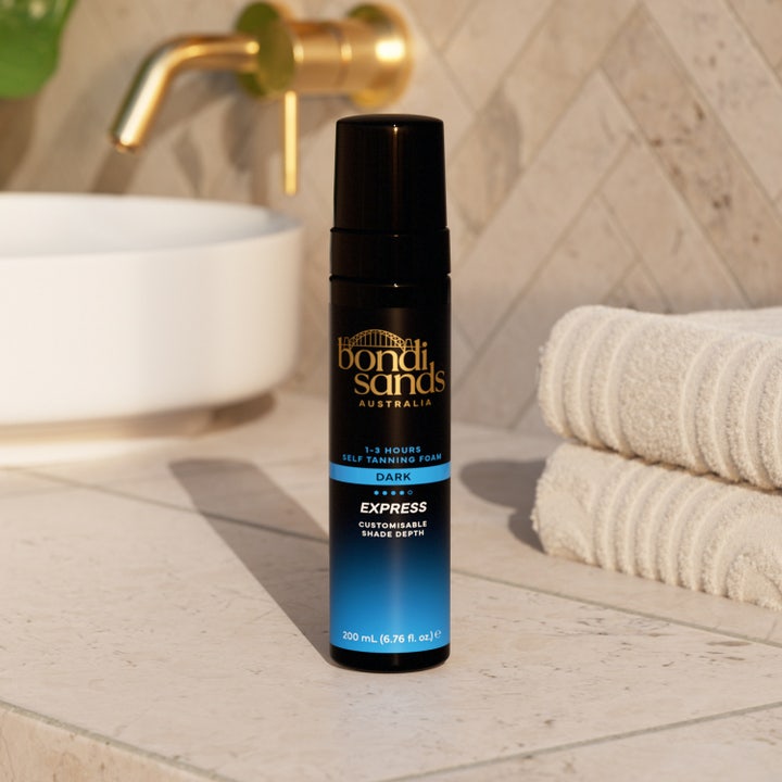 Bondi Sands Self Tan Foam 1 Hour Express 200 ml selvbruning i svart flaske i et lyst baderom med hvite håndklær.
